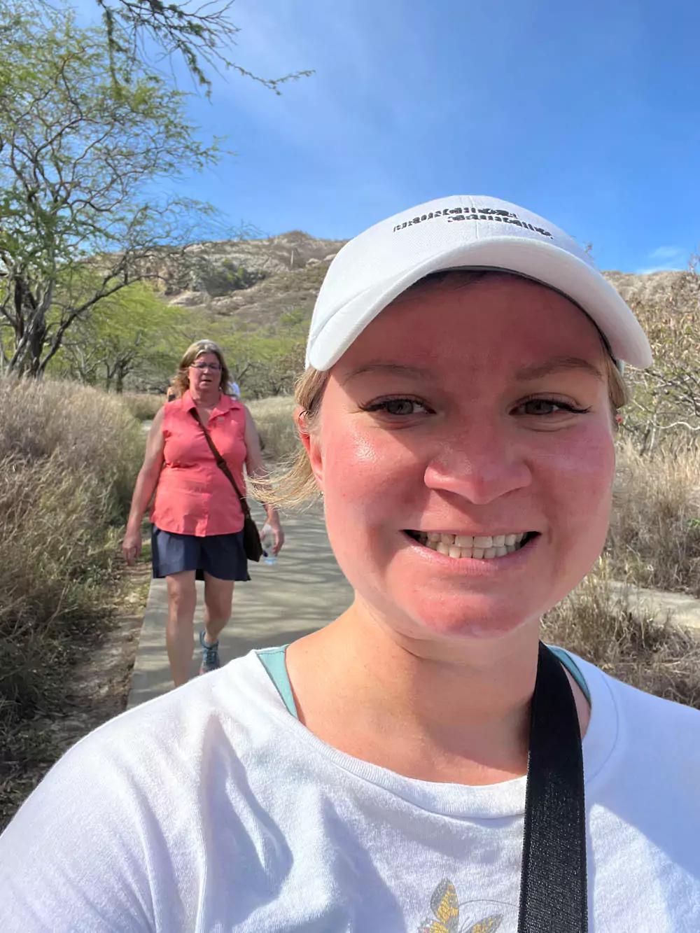 Katie taking a post-hike selfie with Barb in the background at the summit of Diamond Head, both smiling with views of Honolulu and the ocean behind them.