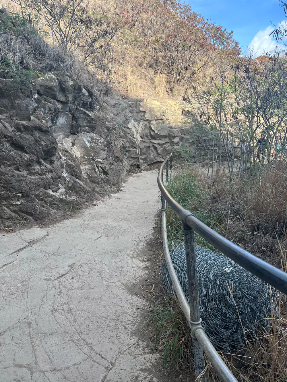 section of the Diamond Head hiking trail on Oʻahu, featuring narrow dirt paths, metal railings, and views of the crater walls under bright tropical sun.