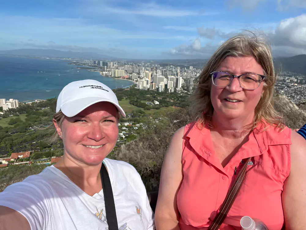 Katie and Barb standing at the summit of Diamond Head State Monument, with windswept hair, scenic views of Honolulu, and the Pacific Ocean stretching out behind them.