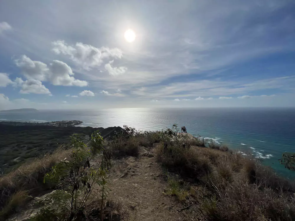 Breathtaking view from the summit of Diamond Head, with the sun shining high above the turquoise Pacific Ocean and waves gently meeting the Oʻahu coastline.