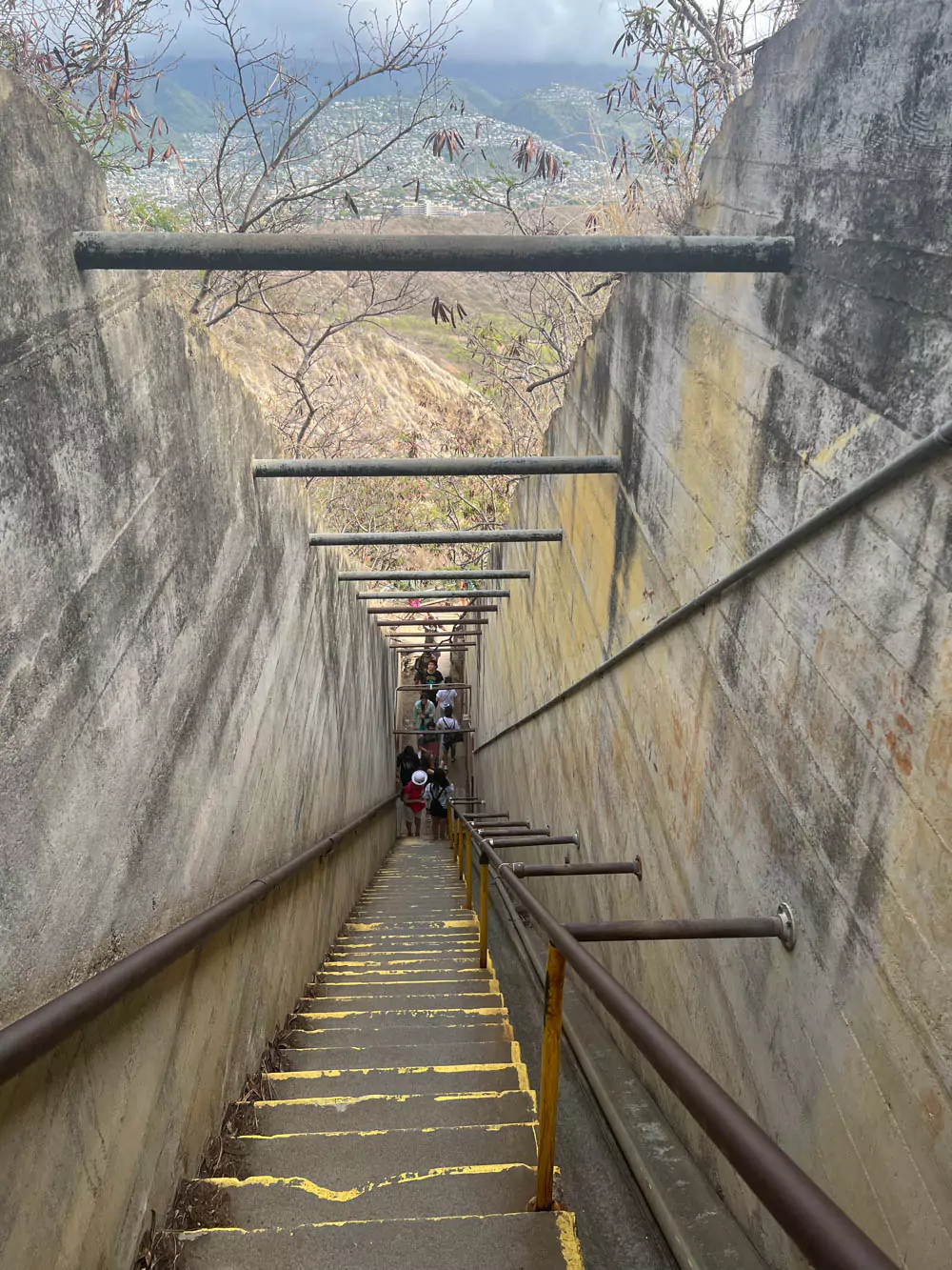 View from the top of the steep staircase on the Diamond Head trail, looking down the narrow steps with handrails and hikers ascending below.