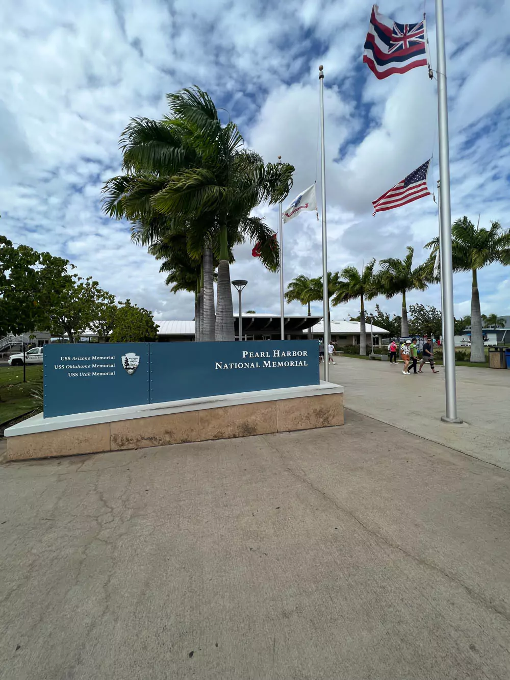 Entrance sign for the Pearl Harbor National Memorial on Oʻahu, Hawaii, featuring the official National Park Service logo and surrounded by palm trees and visitors arriving.