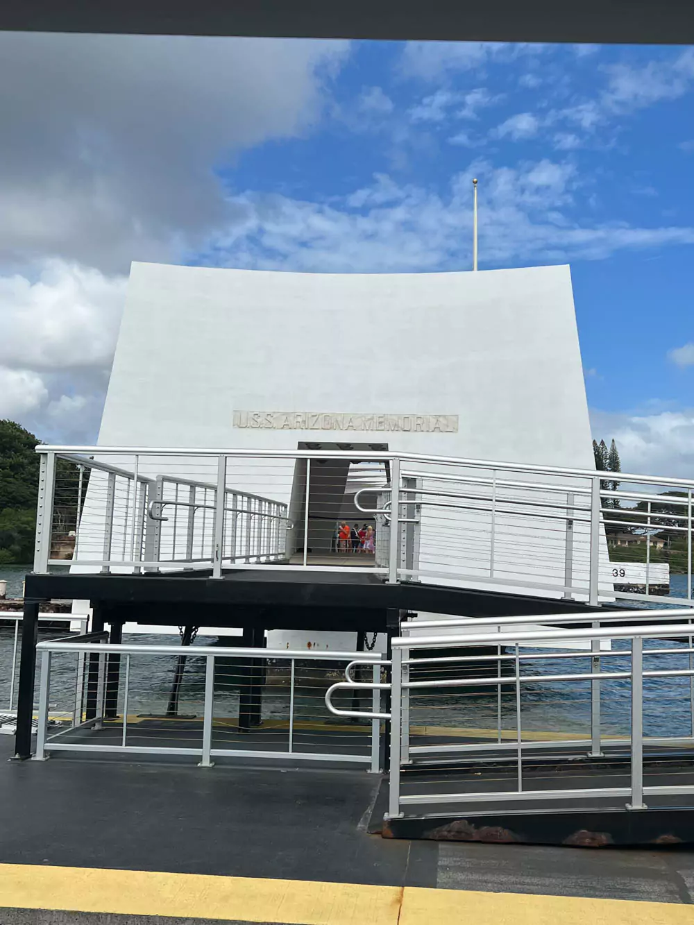 Entrance to the USS Arizona Memorial at Pearl Harbor, showing the sleek white structure with arched openings and American flags flying above as visitors arrive by boat.