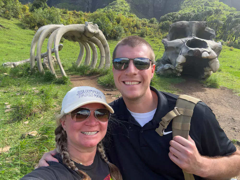Katie and Zack at a Kong: Skull Island filming location in Jurassic Valley at Kualoa Ranch, with towering green cliffs and a movie site sign marking the scene backdrop.