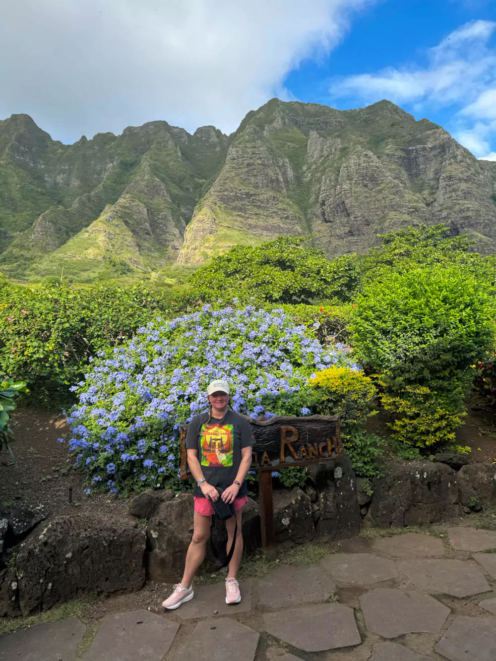 Katie standing in front of vibrant tropical flowers with the lush green Koʻolau Mountains towering behind her at Kualoa Ranch on Oʻahu.