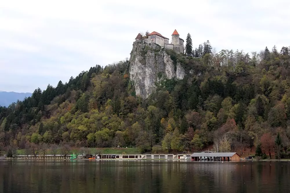 Scenic view of Lake Bled in Slovenia with the medieval Bled Castle perched on a cliff overlooking the turquoise water. The serene lake is surrounded by lush green hills under a clear sky.