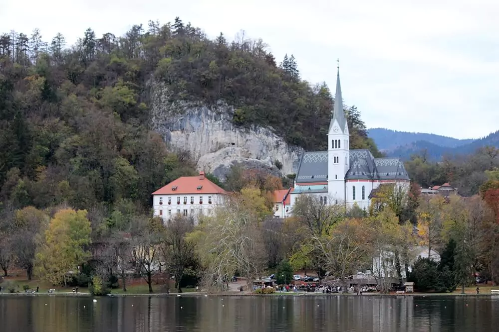 St. Martin Parish Church on Lake Bled, Slovenia, nestled at the foot of a forested hill with its white steeple rising above the trees, framed by the lake’s shoreline and alpine backdrop.