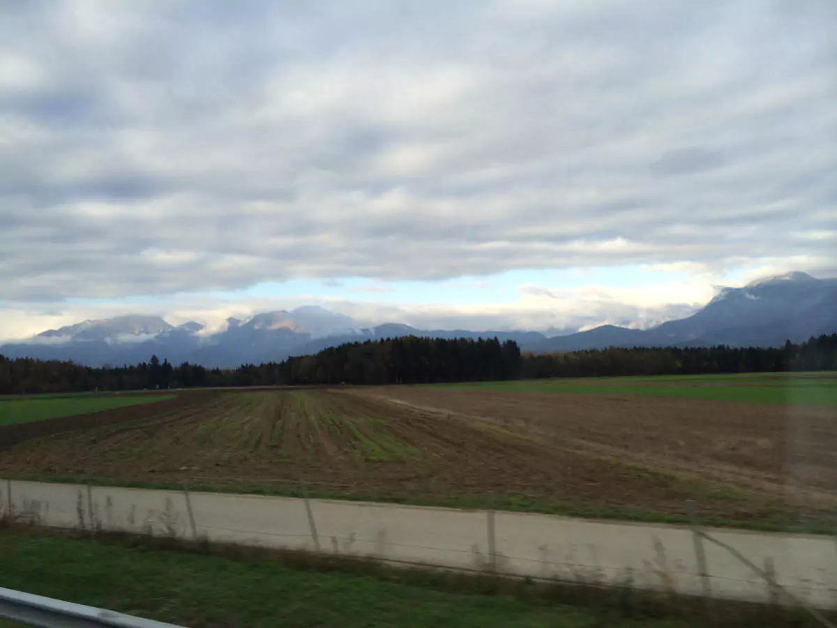 A scenic road winding through the lush green countryside of Slovenia, bordered by fields, trees, and distant hills under a partly cloudy sky. The peaceful rural landscape offers a glimpse of Slovenia’s natural beauty.