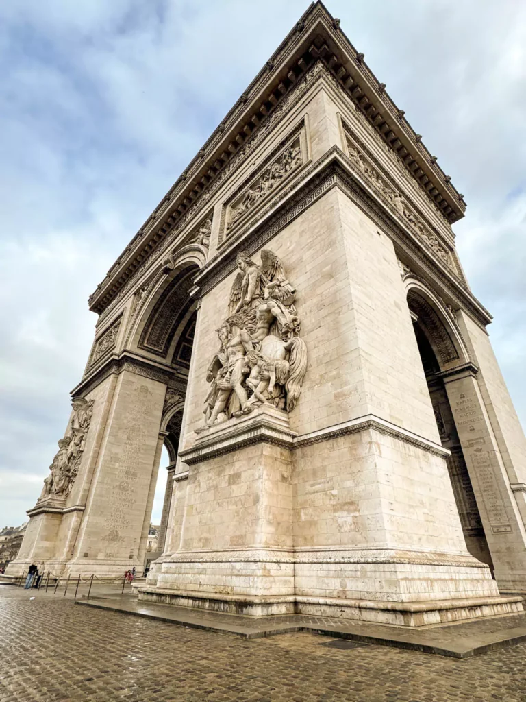 Angled wide-lens side view of the Arc de Triomphe in Paris, France, showcasing the monument’s massive stone arches, intricate sculptural reliefs, and grand scale under a dramatic Parisian sky.