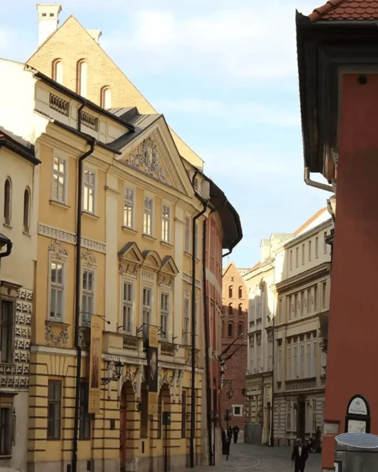 Charming streets in Krakow, Poland, lined with historic buildings featuring colorful facades and traditional architecture. Pedestrians walk along cobblestone roads under clear skies, capturing the city’s vibrant urban life.