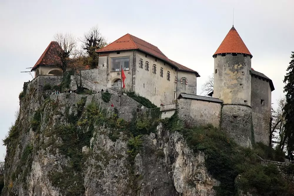 Bled Castle perched on a steep cliff overlooking Lake Bled in Slovenia, featuring red rooftops, stone walls, and panoramic views of the lake and surrounding Julian Alps.