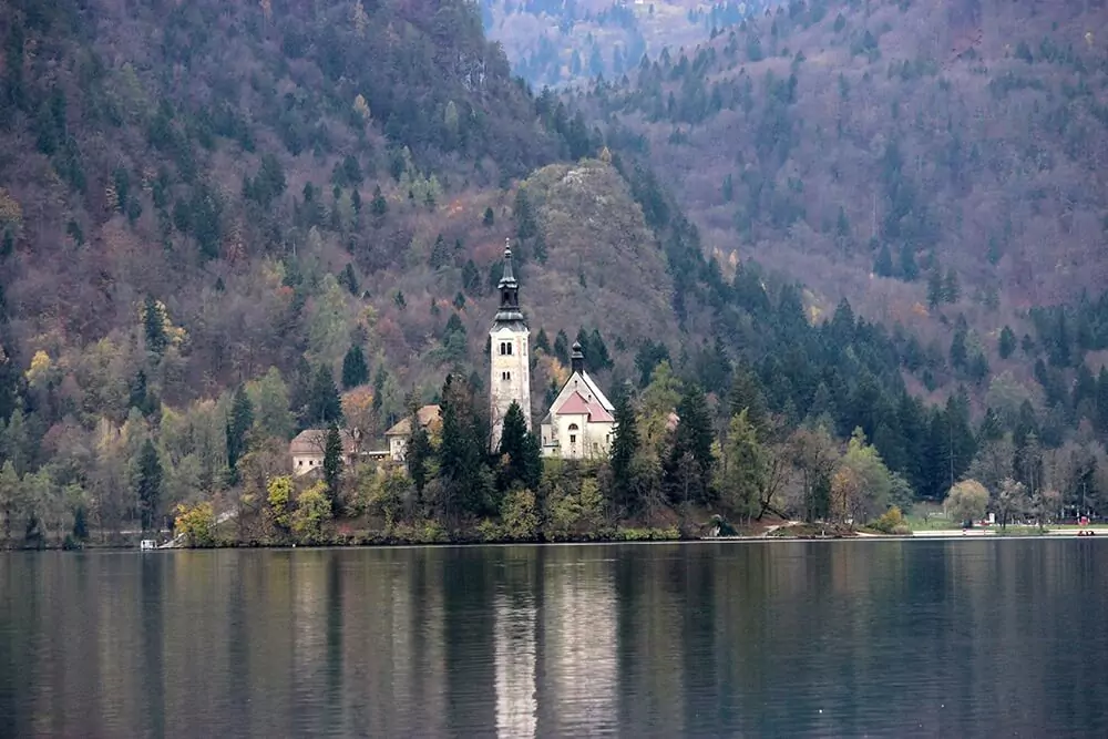 Small island on Lake Bled, Slovenia, featuring the Church of the Assumption with its bell tower surrounded by trees, set in turquoise waters with mountains in the distance.