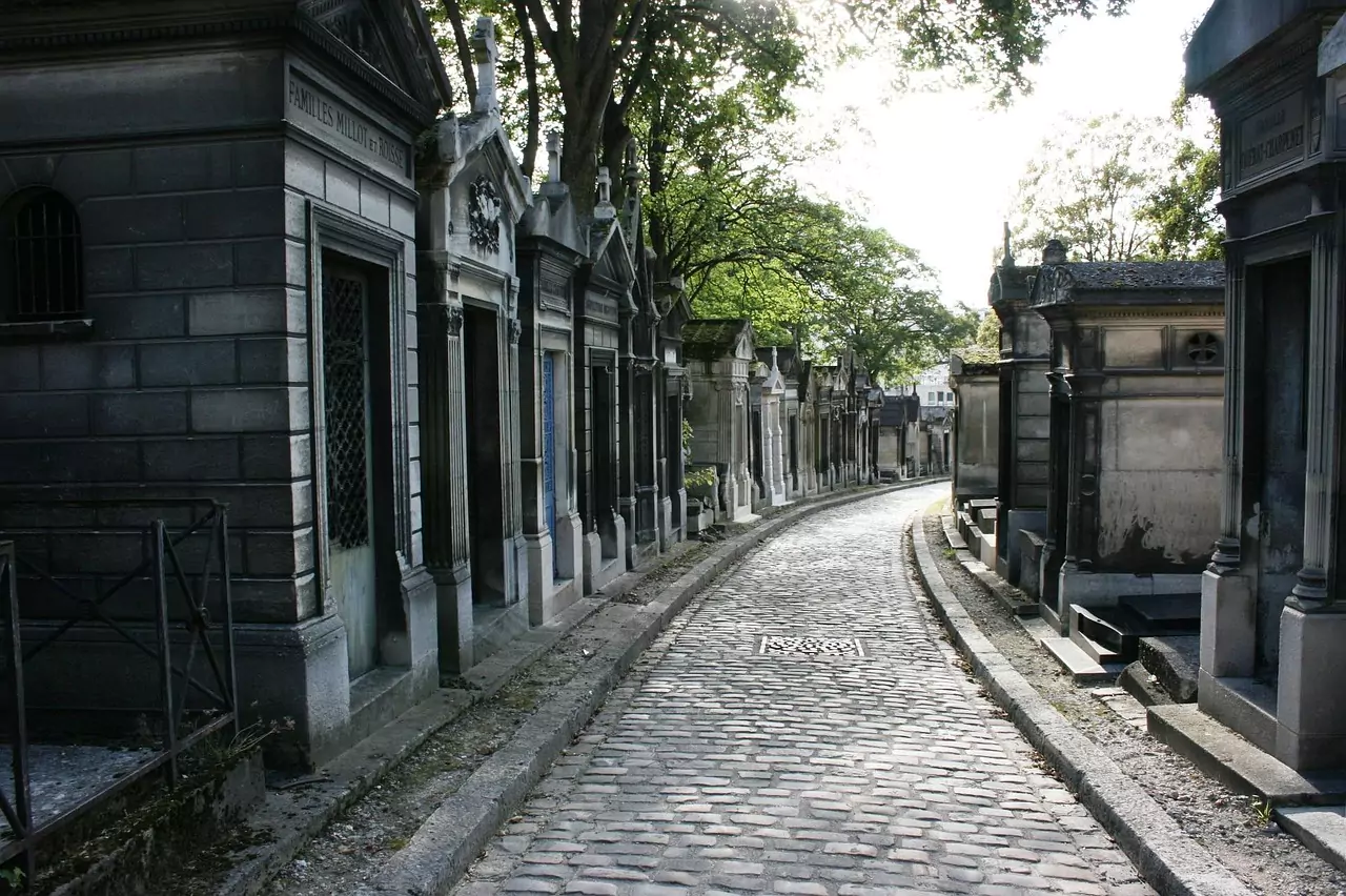 Cobblestone street winding through Père Lachaise Cemetery in Paris, lined with historic mausoleums, tombstones, and tall trees casting dappled shadows along the quiet pathway.