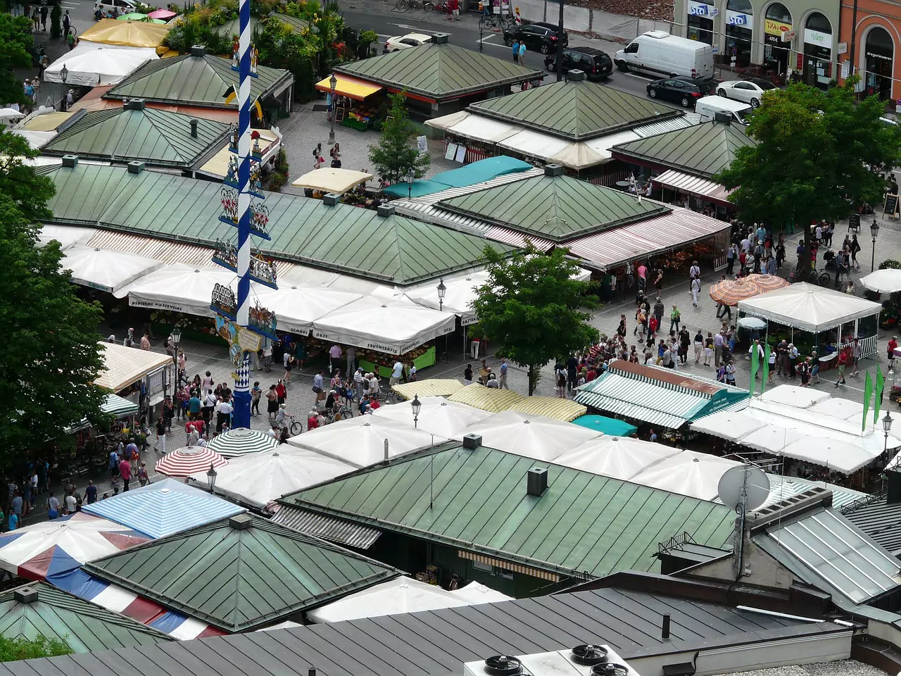 Bird’s-eye view of Viktualienmarkt, Munich’s famous open-air market, showing colorful stalls arranged in a bustling plaza surrounded by historic buildings. Shoppers and vendors create a lively atmosphere in this central city destination.