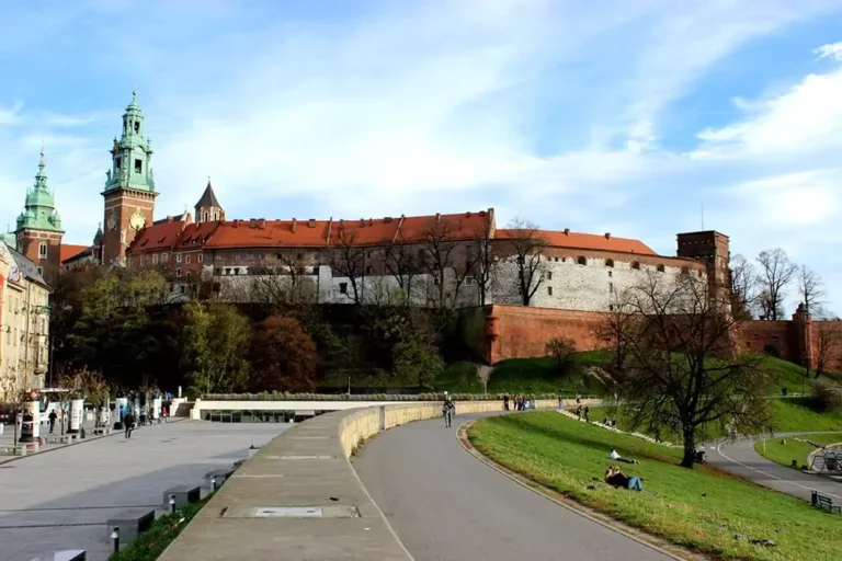 Street-level view of Wawel Castle in Kraków, Poland, seen from a nearby sidewalk with castle towers rising above red-brick walls and trees lining the foreground.