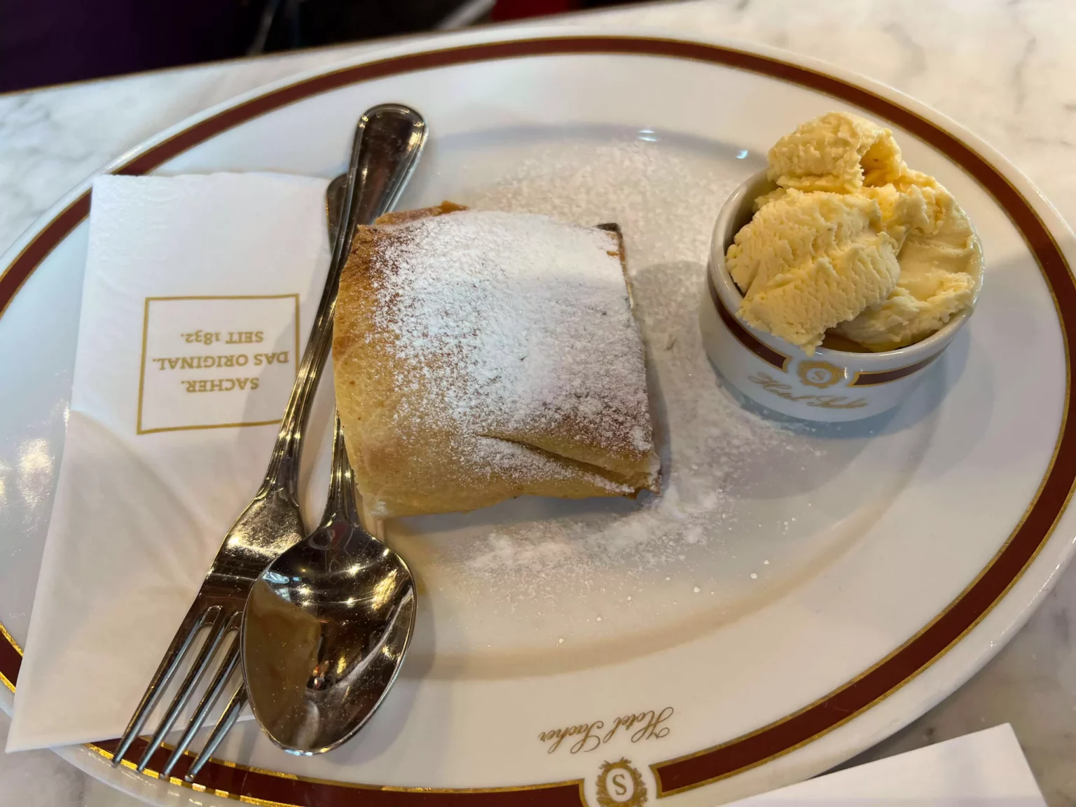 A plate of warm apple strudel served with a scoop of vanilla ice cream at Café Sacher in Vienna, Austria. The flaky pastry, dusted with powdered sugar, pairs perfectly with the creamy ice cream in this classic Austrian dessert presentation.