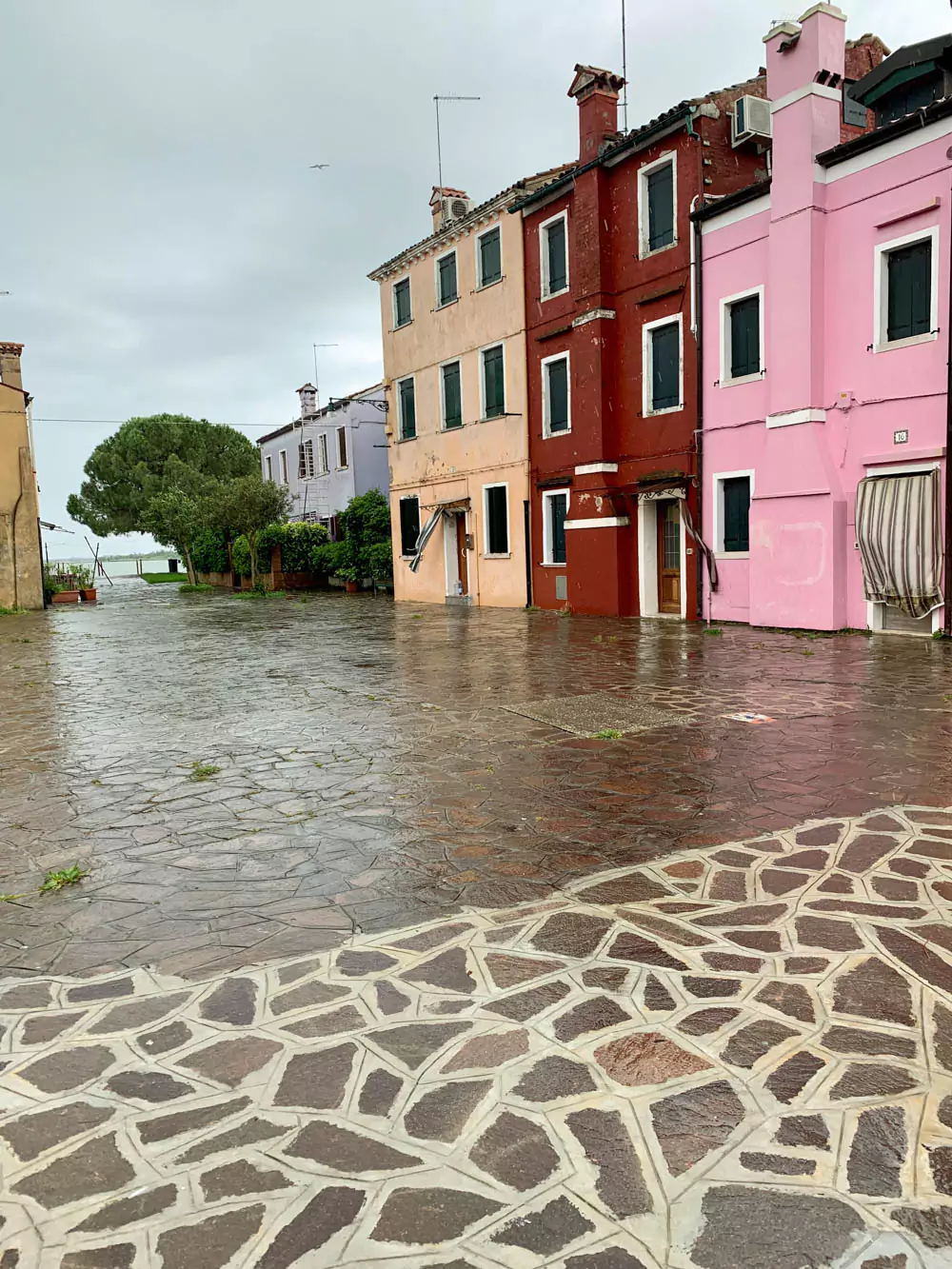A row of vividly painted houses in Burano, Italy, featuring bright pink and orange facades along a narrow street with rain-slicked cobblestones, capturing the island’s famous cheerful and colorful charm.
