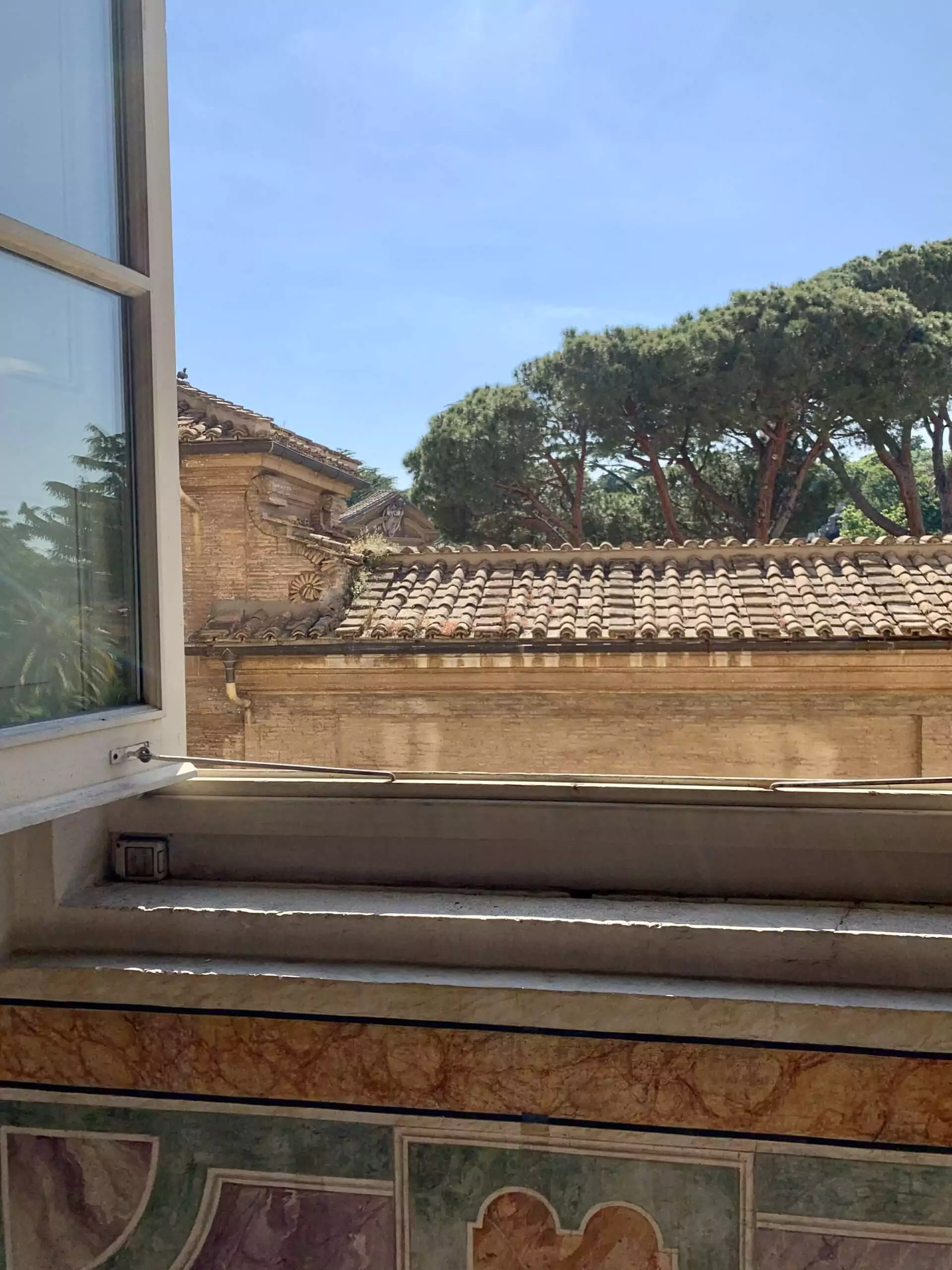 View from a Vatican Museums window looking out at a tall Italian cypress tree and terracotta-tiled rooftops, capturing the classic Roman landscape bathed in natural light.
