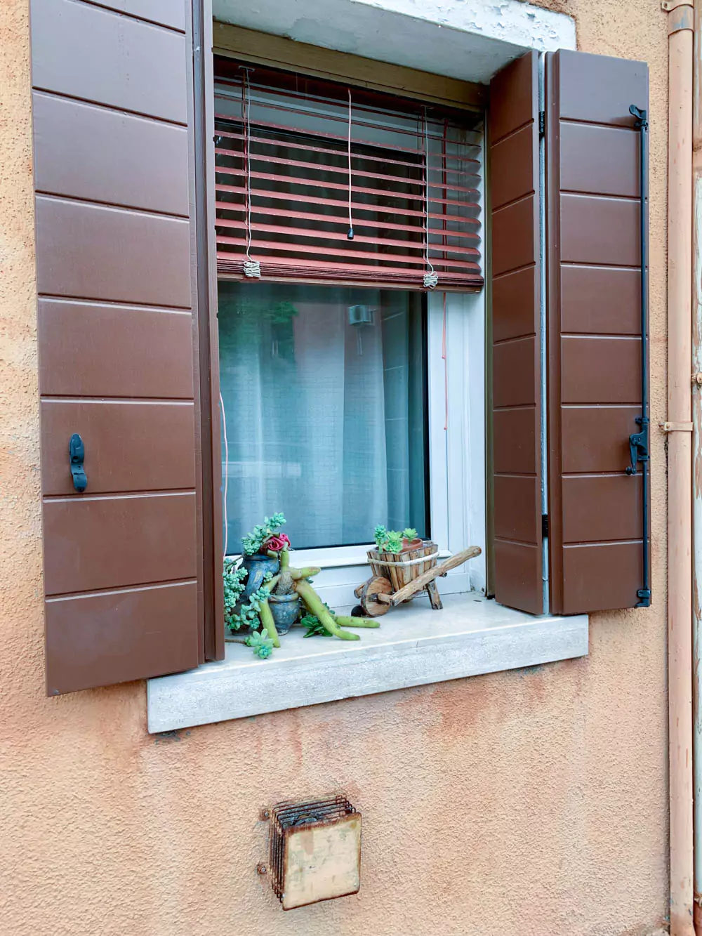 A charming window sill in Burano, Italy, features a miniature wheelbarrow planter filled with vibrant flowers. The playful detail adds a touch of whimsy against the colorful stucco wall, capturing the island’s cozy, artistic spirit.