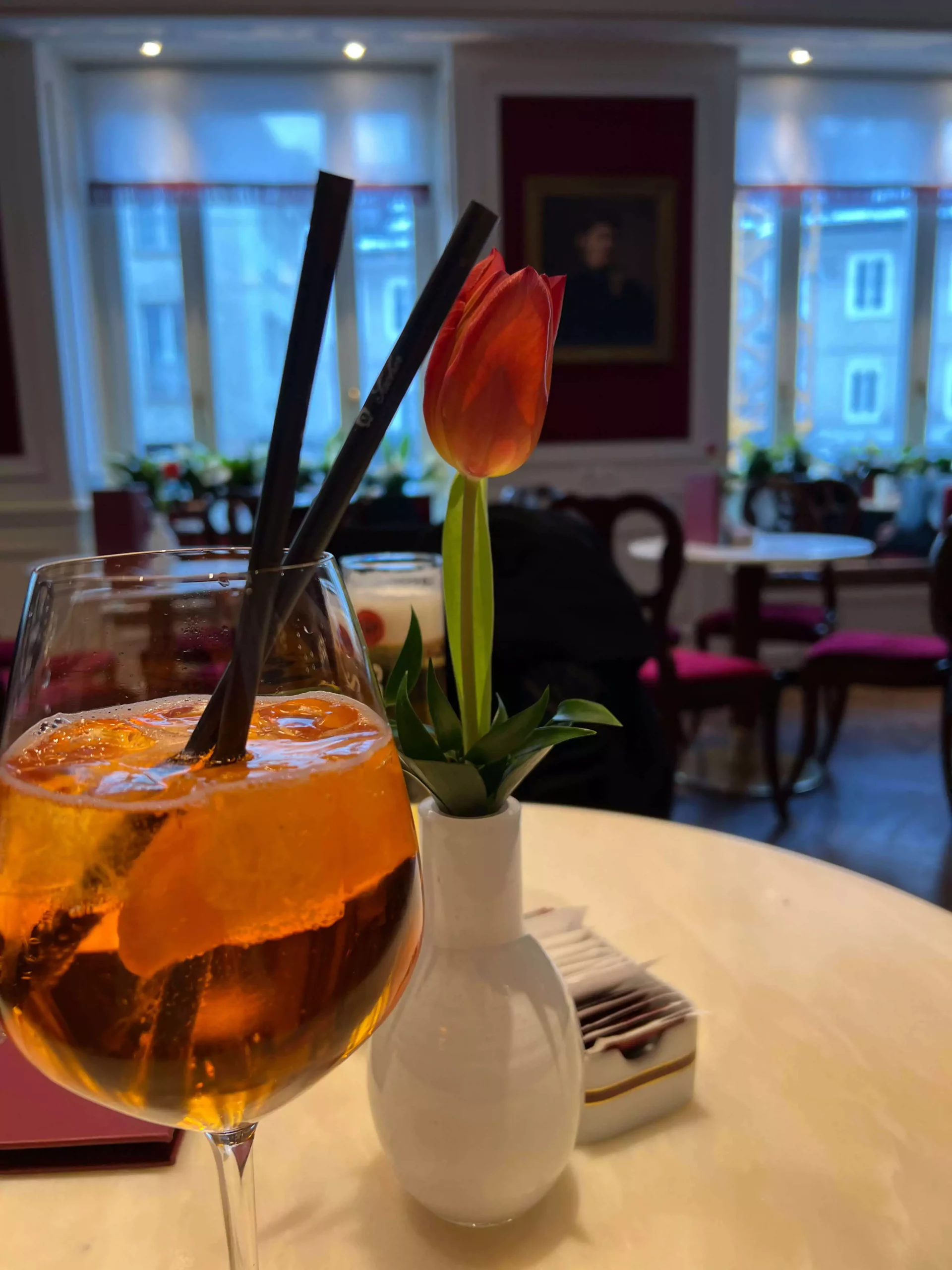 An Aperol Spritz sits on a table inside Café Sacher in Salzburg, Austria, glowing with its signature orange hue. The elegant glassware and classic interior add to the refined, cozy atmosphere of the café.