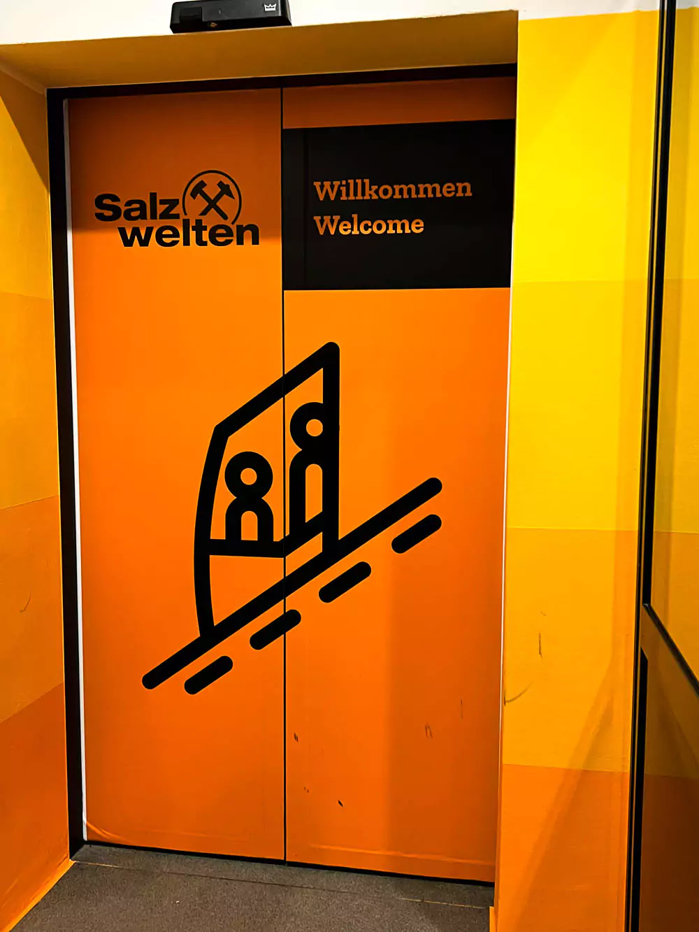 Bright orange door leading to the funicular room at the Hallstatt Salt Mine (Salzwelten), with industrial-style metal framing and warning signs. The vivid color adds visibility and marks the transition to the steep mountain railway.
