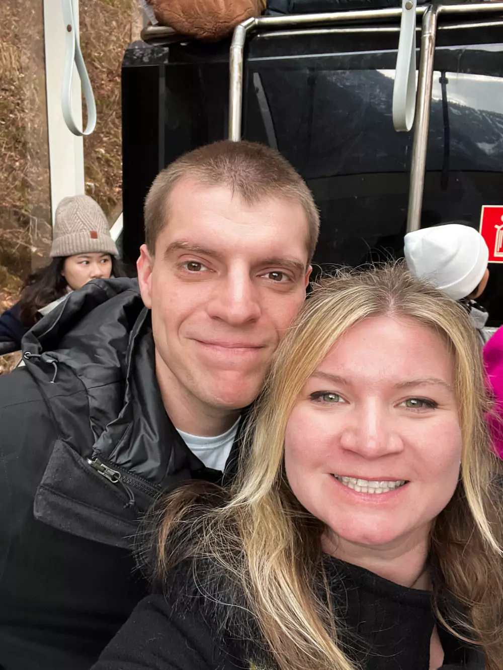Katie and Zack taking a selfie inside the funicular at Hallstatt Salt Mine (Salzwelten), smiling with the steep forested hillside visible through the window behind them. The cozy ride marks the start of their scenic alpine adventure.