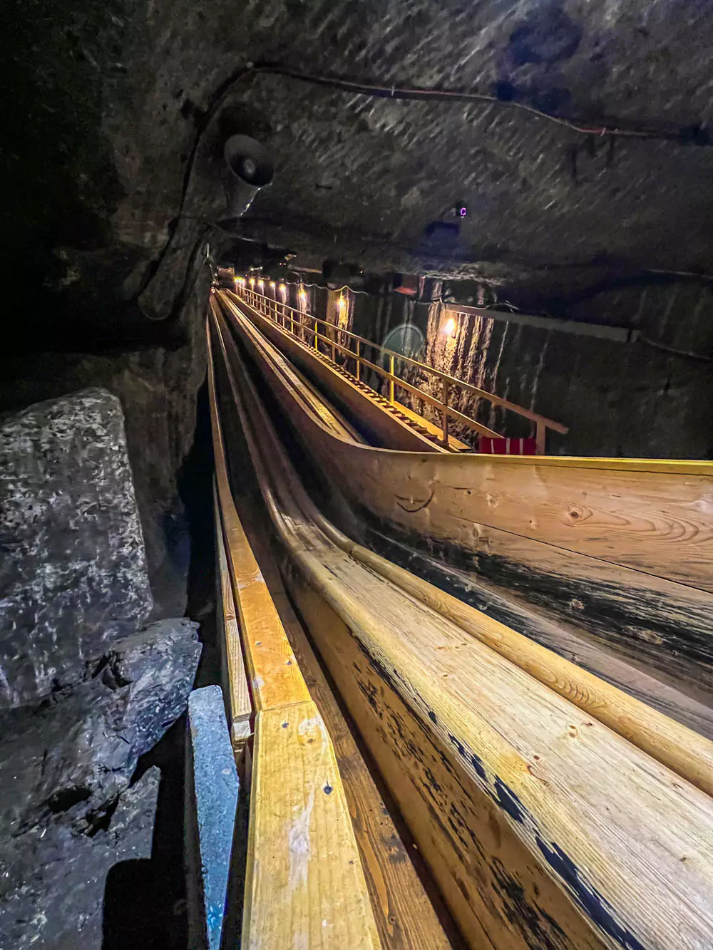 Second wooden slide inside Hallstatt Salt Mine (Salzwelten), longer and steeper, with a shiny, worn surface from repeated use. The slide is lit by soft overhead lighting and surrounded by rugged tunnel walls, offering an adventurous descent into the mine.