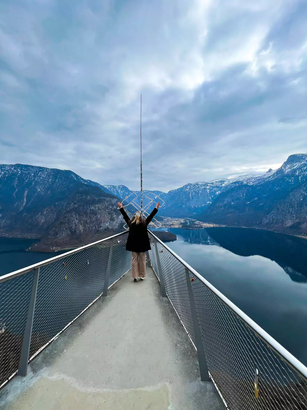 Katie standing at the top of the Welterbeblick Skywalk in Hallstatt, Austria, smiling with a panoramic view of the village, lake, and surrounding mountains stretching out behind her.