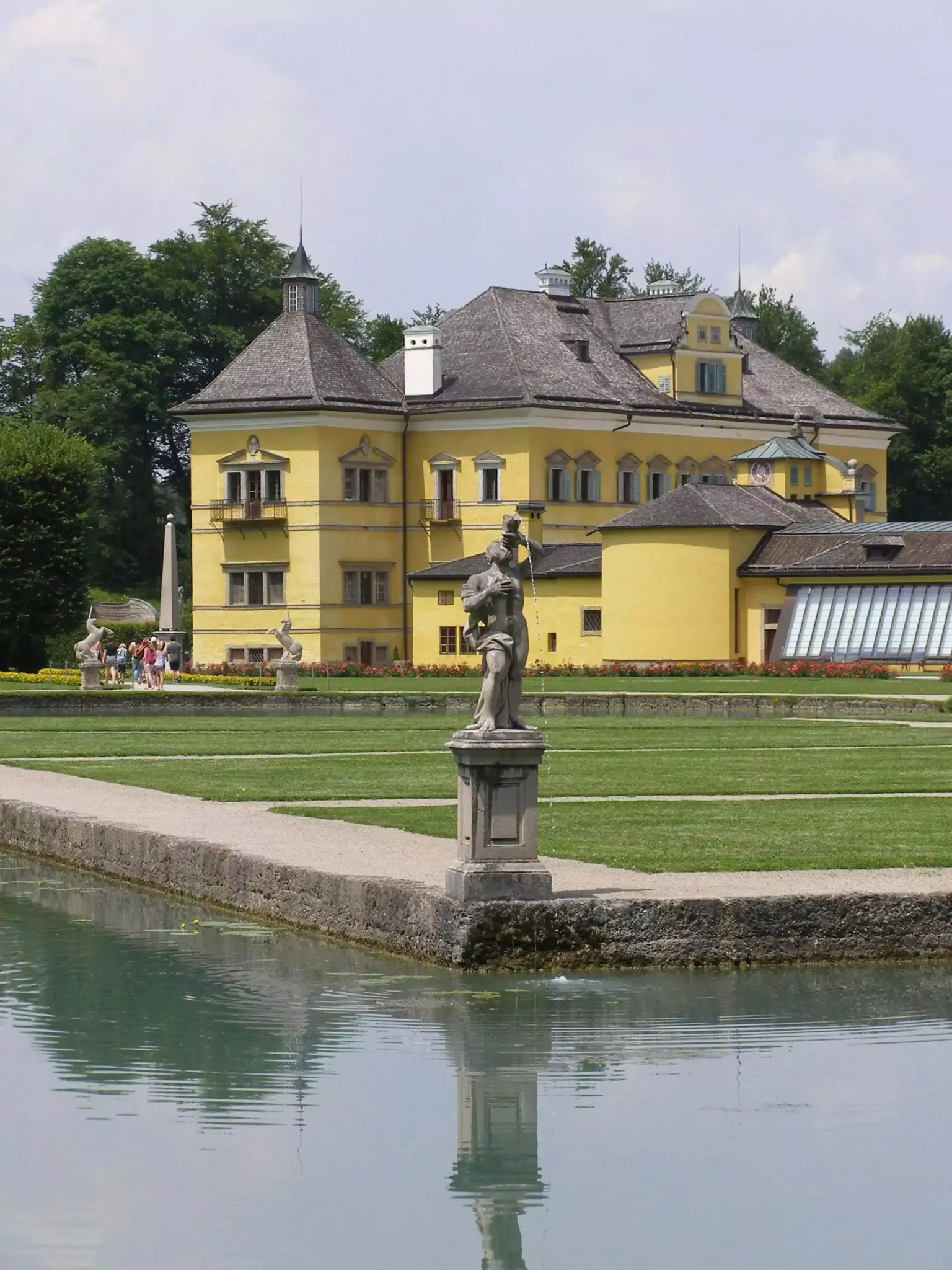Exterior of Schloss Hellbrunn in Salzburg, Austria, featuring a vibrant yellow Baroque facade with green shutters, set against a backdrop of landscaped grounds and wooded hills.