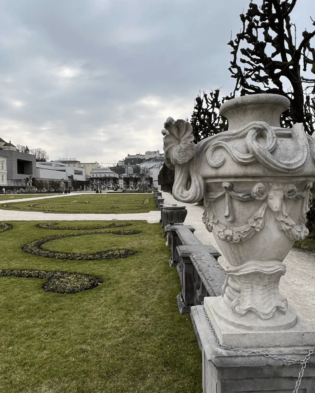 Ornate stone vases lining the manicured lawn of Schloss Mirabell gardens in Salzburg, Austria, symmetrically placed along gravel paths with flowerbeds and trimmed greenery.