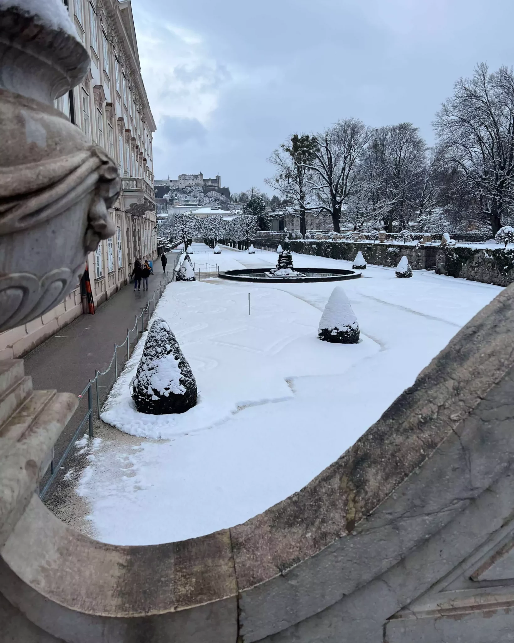 Snow-dusted Mirabell Palace Gardens in Salzburg, Austria, featuring neatly landscaped paths, classical statues, and a view of the Baroque palace in the background.