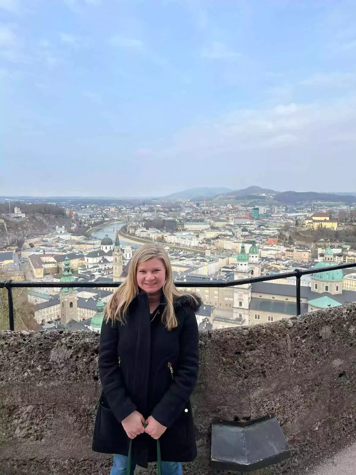 Katie standing at the Hohensalzburg Fortress, overlooking a panoramic view of Salzburg, Austria, with the city’s rooftops, church spires, and distant mountains visible beneath a bright sky.