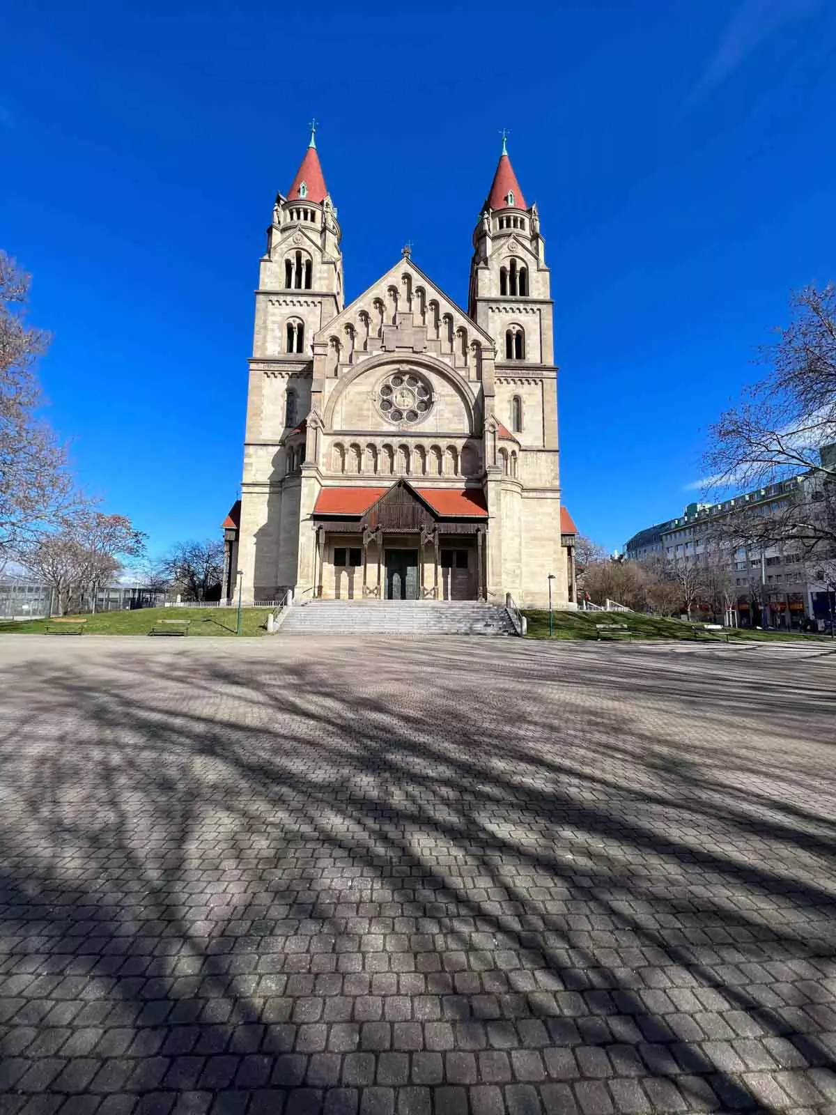Exterior of Kirche St. Franziskus von Assisi in Vienna, Austria, featuring striking red-tiled towers, Romanesque Revival design, and arched entrances, set along the Danube River in the Mexikoplatz district.