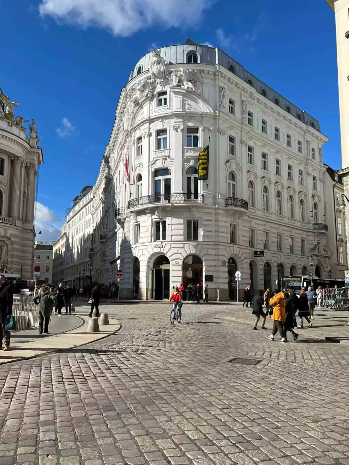 Cobblestone streets of Michaelerplatz in Vienna, Austria, curving around the historic square with horse-drawn carriages, elegant buildings, and the grand entrance to Hofburg Palace in view.