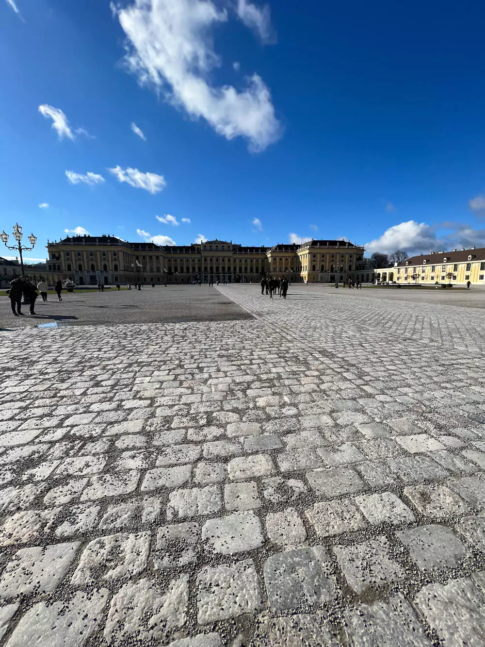 Entrance to Schloss Schönbrunn in Vienna, Austria, featuring ornate wrought-iron gates, golden eagle statues, and a wide plaza leading toward the majestic yellow Baroque palace in the distance.