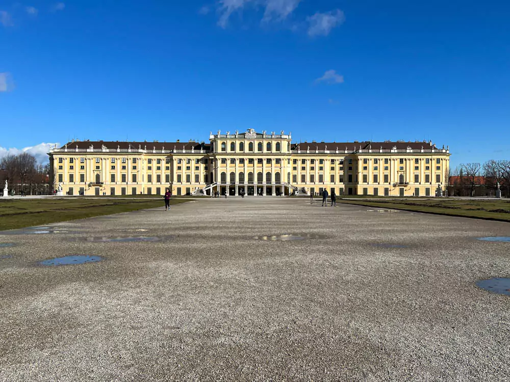 South façade of Schönbrunn Palace in Vienna, Austria, during winter, with the grand yellow Baroque building framed by snow-dusted grounds and bare trees under a pale sky.