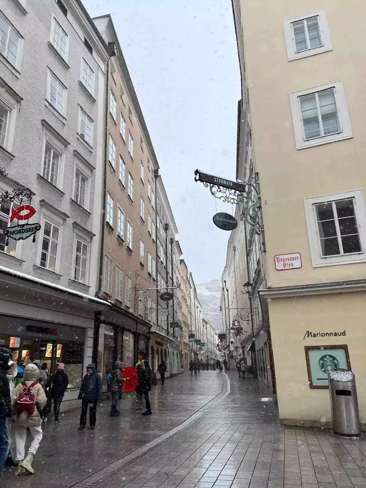 Snow-covered street in Salzburg’s historic city centre, with baroque buildings, church spires, and pedestrians walking through the quiet, wintry atmosphere beneath softly falling snow.