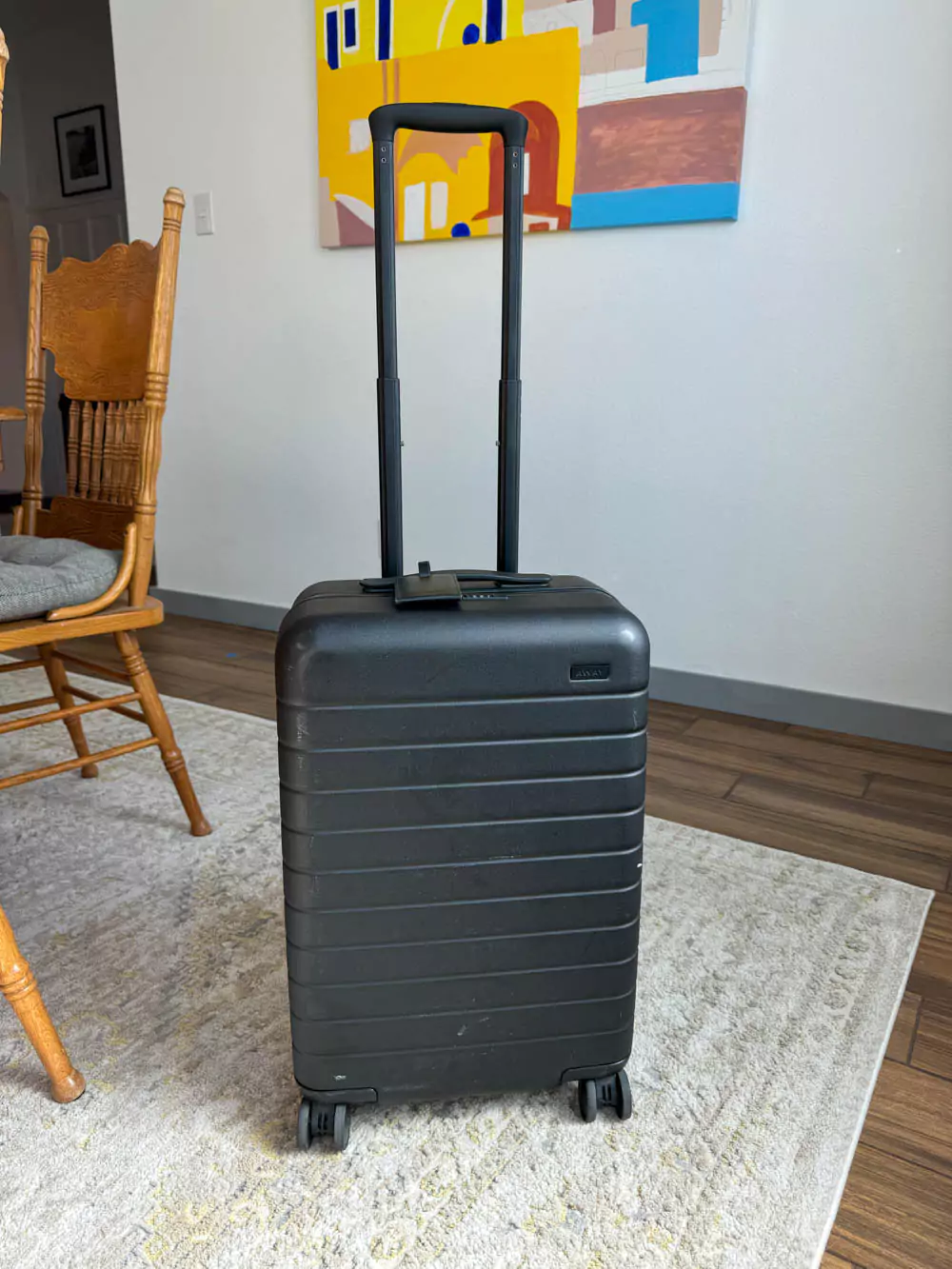 AWAY Bigger Carry-On Suitcase in black, standing in a dining room with hardwood floors and natural light, showcasing its sleek design and durable shell — a stylish travel essential