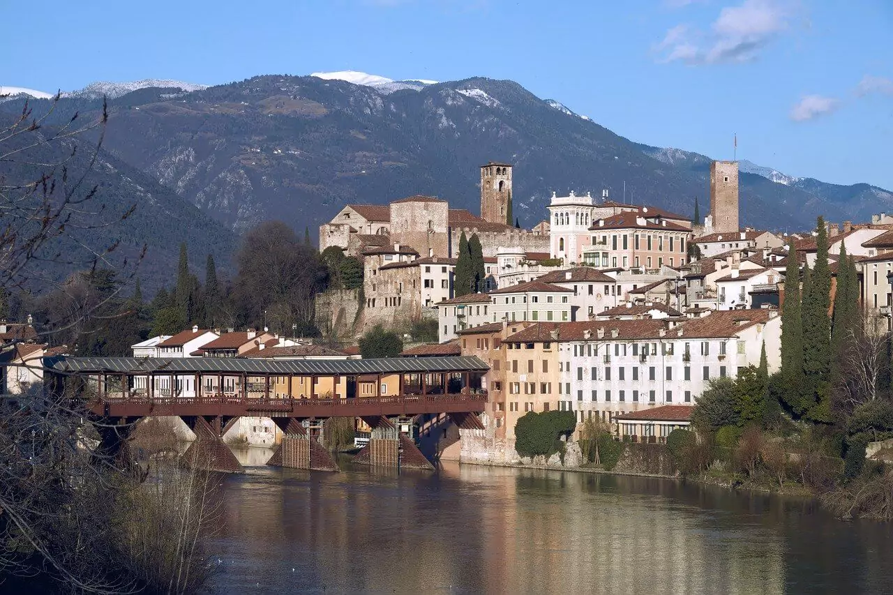 Bassano del Grappa, Italy, with a view of the historic wooden Ponte Vecchio bridge spanning the Brenta River, framed by colorful buildings and alpine foothills 