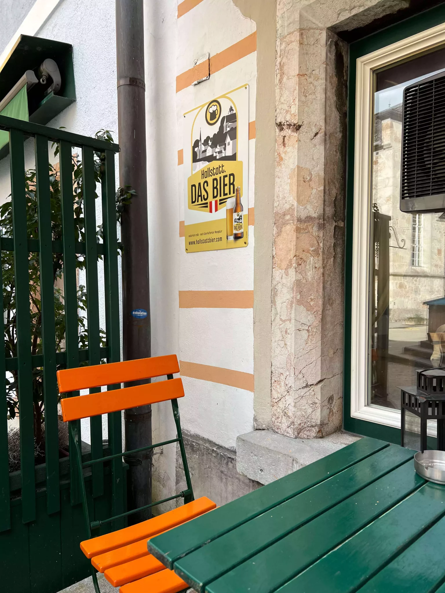 A beer sign hangs on the exterior of a restaurant in Hallstatt, Austria, above a cozy setup with an orange chair and a green table. The colorful furniture and rustic charm create a welcoming alpine atmosphere for outdoor dining.