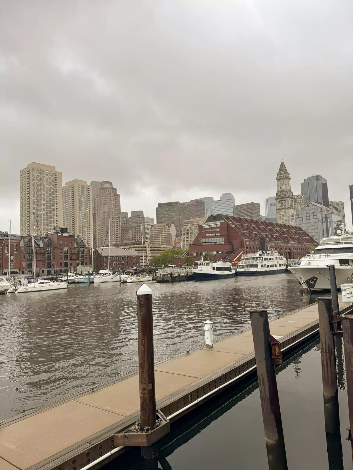 Cloudy, rain-soaked view of Boston Harbor with the city skyline in the background, featuring misty high-rises and choppy water under a gray sky. The moody atmosphere captures the coastal city's quiet, stormy side.