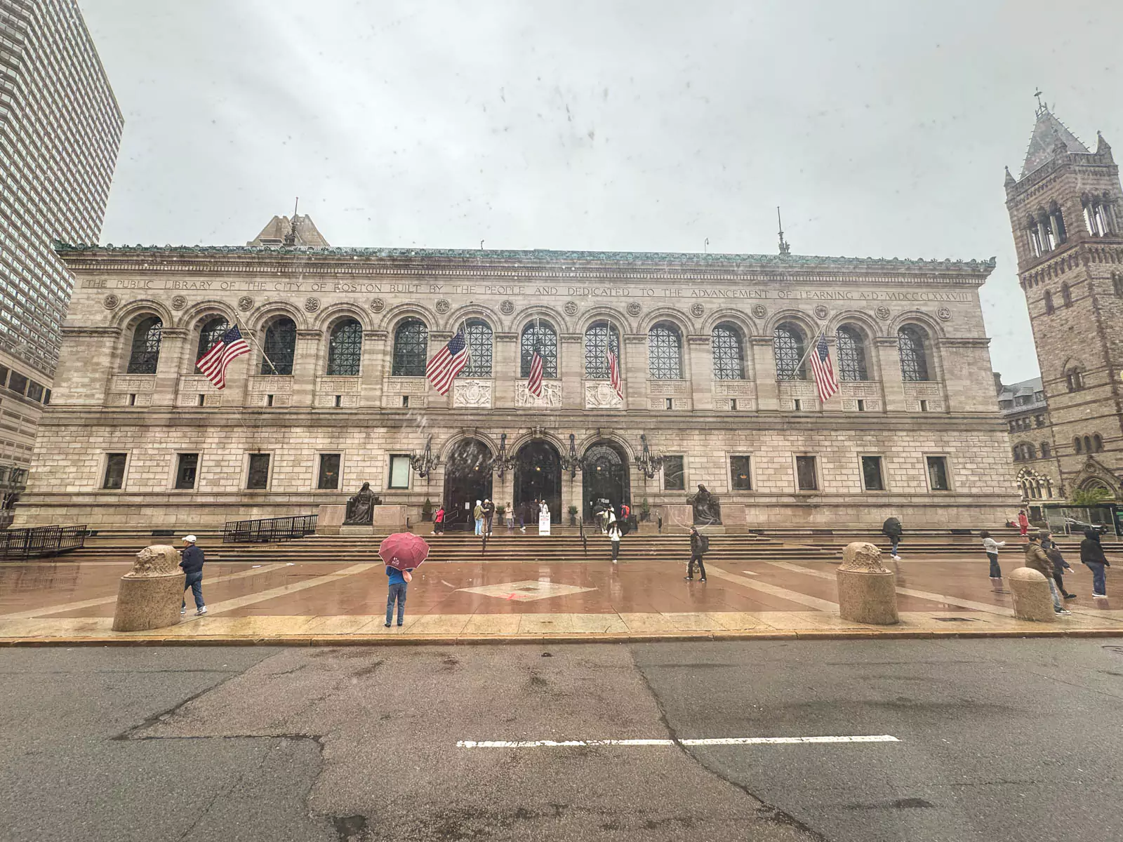 Exterior of the Boston Public Library on a rainy day, with wet pavement reflecting the building’s grand Beaux-Arts architecture. Umbrella-covered visitors pass by the arched windows and stone facade, adding a cozy, atmospheric feel.