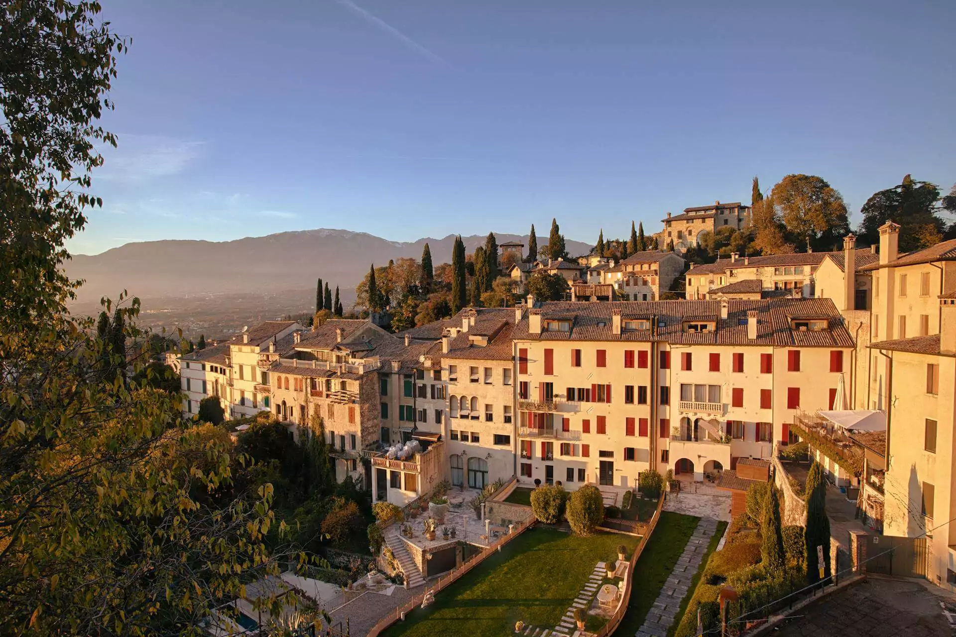 Brown and white concrete buildings in Asolo, Italy, surrounded by manicured green lawns and cypress trees, with distant views of rolling hills