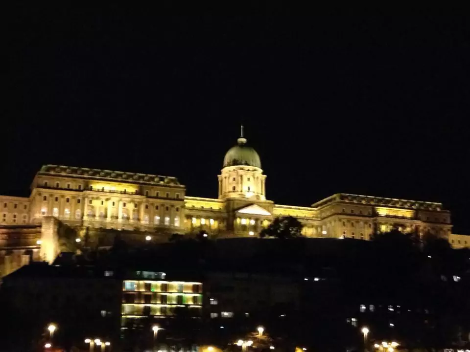 Buda Castle illuminated against the dark night sky in Budapest, with its grand facade glowing warmly and reflections visible in the Danube River below. The historic castle stands as a stunning nighttime landmark overlooking the city.