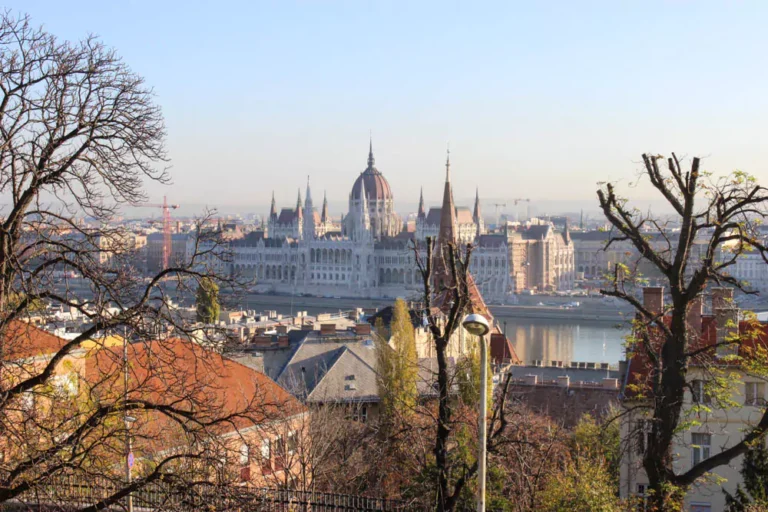 The Hungarian Parliament Building in Budapest bathed in daylight, displaying its ornate Gothic Revival architecture with red-tiled roofs and tall spires. The Danube River flows in front, with clear skies enhancing the grandeur of this iconic landmark.