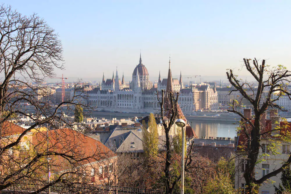 The Hungarian Parliament Building in Budapest bathed in daylight, displaying its ornate Gothic Revival architecture with red-tiled roofs and tall spires. The Danube River flows in front, with clear skies enhancing the grandeur of this iconic landmark.