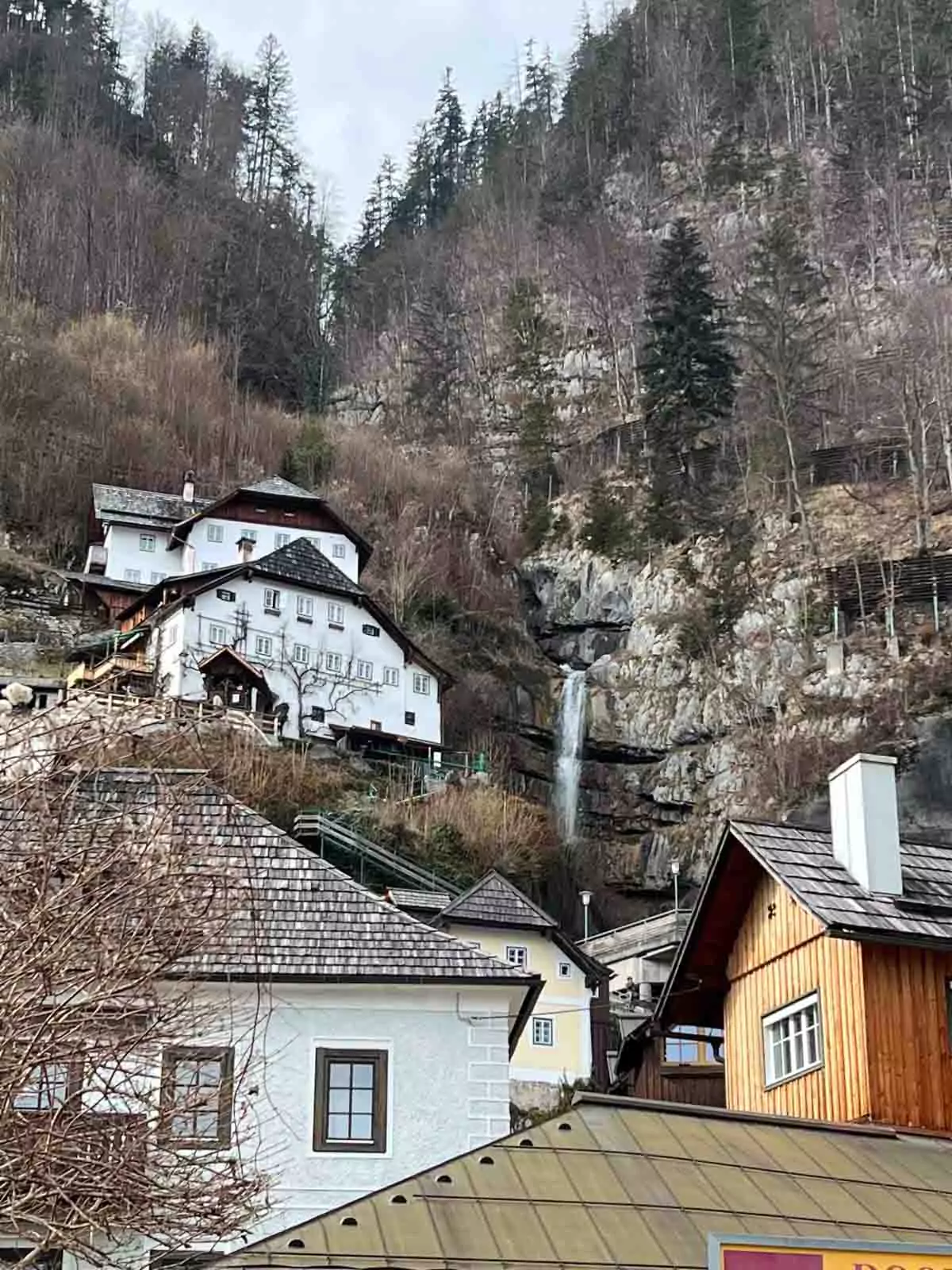 Charming alpine buildings nestled beside a cascading waterfall in Hallstatt, Austria, with traditional wooden facades and balconies surrounded by lush greenery and dramatic mountain cliffs.