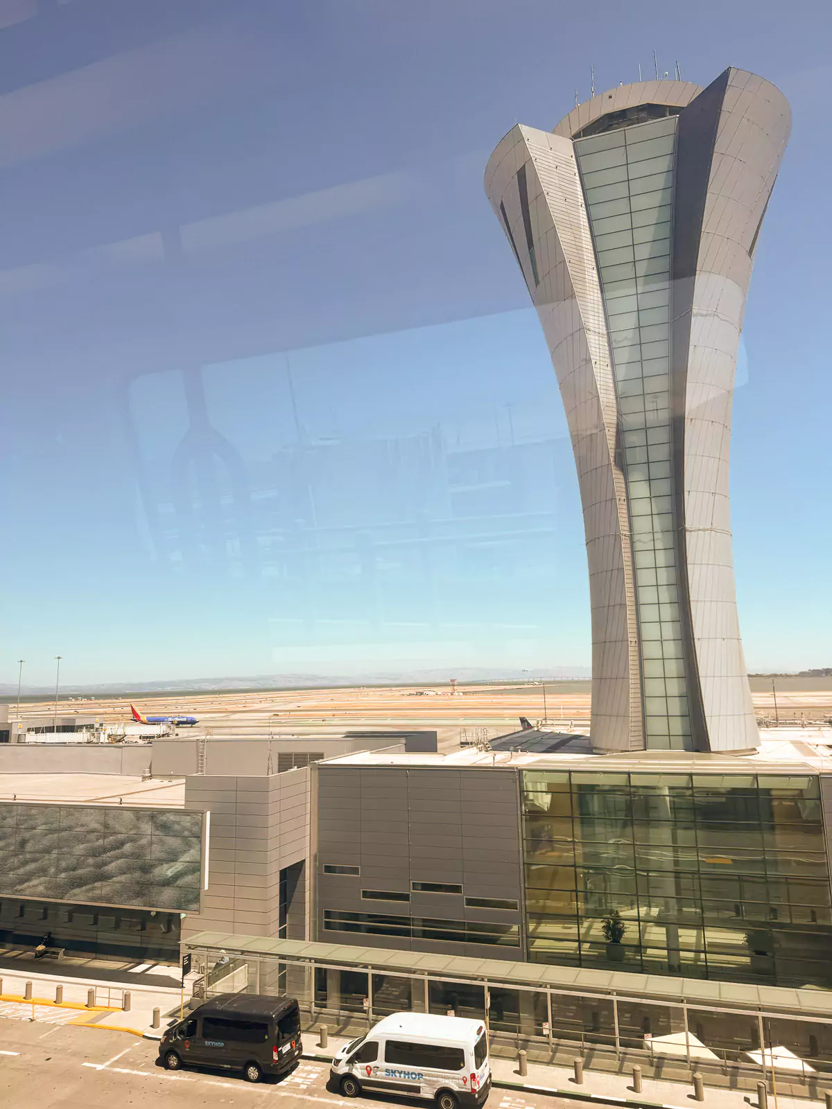 The San Francisco Airport control tower stands tall as an airplane descends for landing nearby. The busy airport scene shows clear skies and runway activity.
