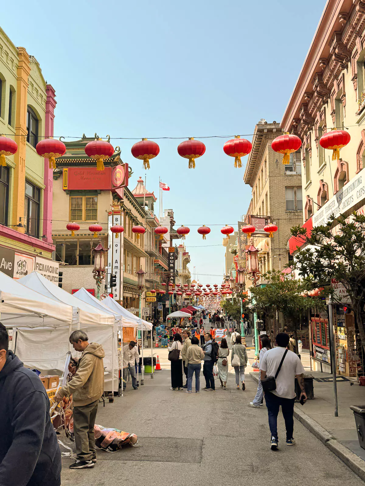A busy street in San Francisco’s Chinatown with hanging lanterns, Chinese shop signs, and people walking. The street is lined with stores and has a lively city atmosphere.