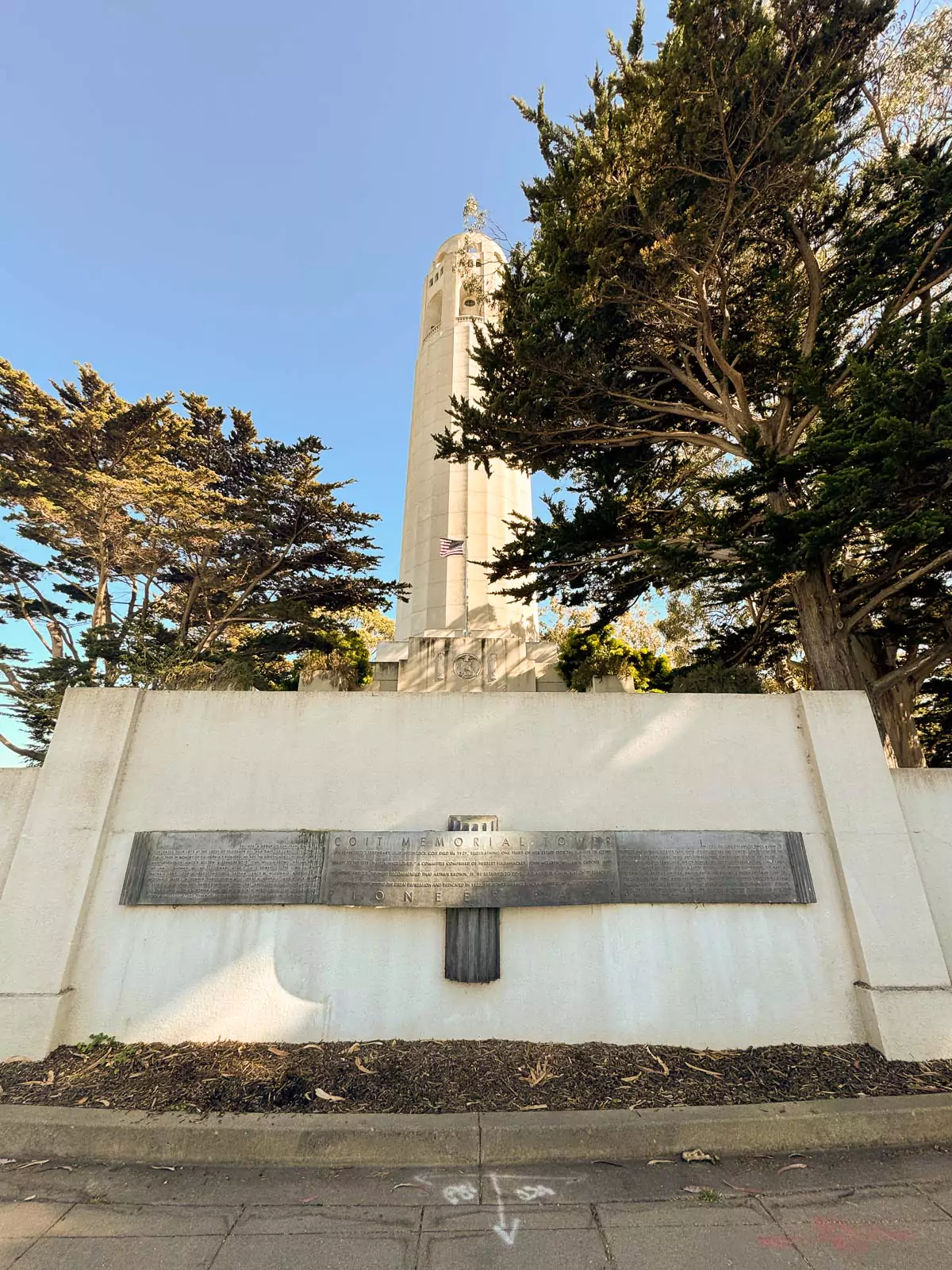 View from the base looking up at Coit Tower in San Francisco, with its white cylindrical structure rising against a clear blue sky. The perspective emphasizes the tower’s height and iconic status on Telegraph Hill.